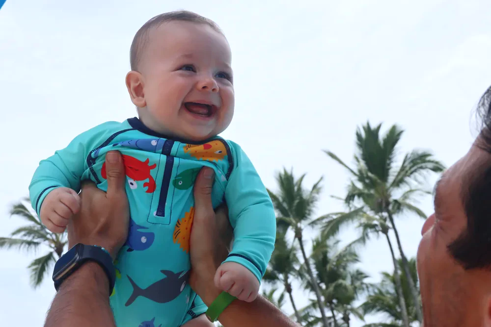 Bebê sorrindo com roupa Pulpo enquanto é erguido por um adulto ao ar livre
